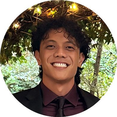 cameron-headshot A person in a dark suit and tie smiles at the camera while standing under foliage with string lights.