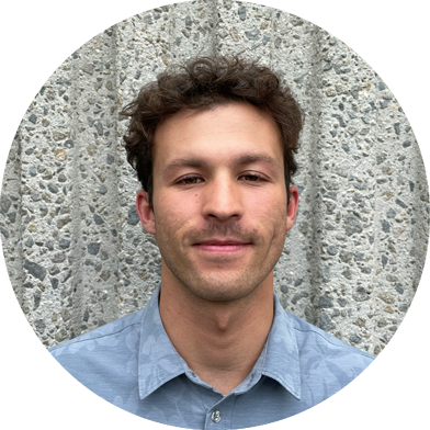 nao-headshots Young man with short curly brown hair and a mustache, wearing a light blue collared shirt, standing in front of a textured concrete wall.
